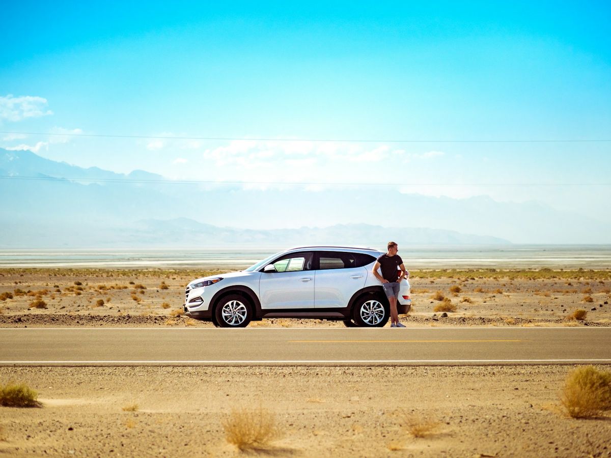 Man standing beside a white SUV on a road under blue sky